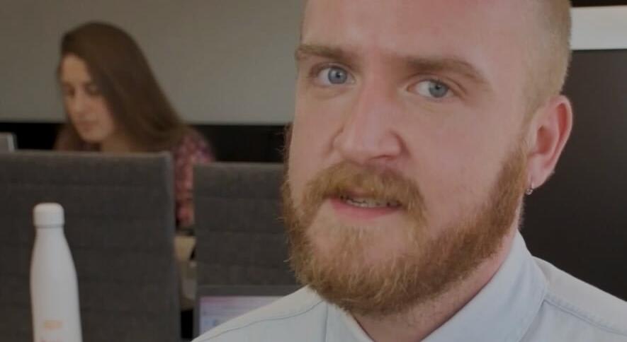 bearded man in light blue shirt, focused in modern office backdrop with woman working, professional workspace atmosphere