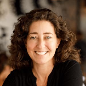 smiling woman with curly hair in a casual black shirt, natural light background with a warm, inviting atmosphere