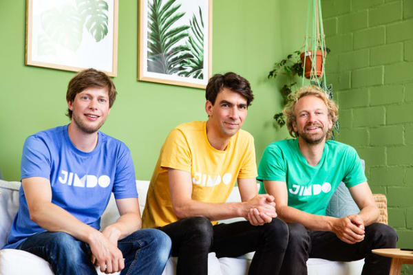 JIMDO team members in colorful shirts, posing against a green background with plants and framed artworks., JIMDO, team_photo