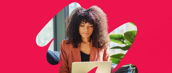 woman with curly hair using a laptop in a modern, bright setting with green plants and a bold pink background, focused woman working on laptop in stylish workspace with plants and vibrant colors