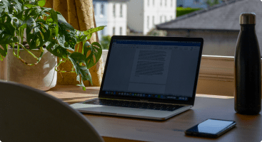 laptop-on-wooden-desk-with-notepad-and-phone, sunlit-window-view-with-green-plant-and-thermal-water-bottle