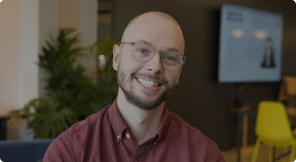 smiling man with glasses in casual attire against a modern office backdrop, green plants and digital screen visible in background
